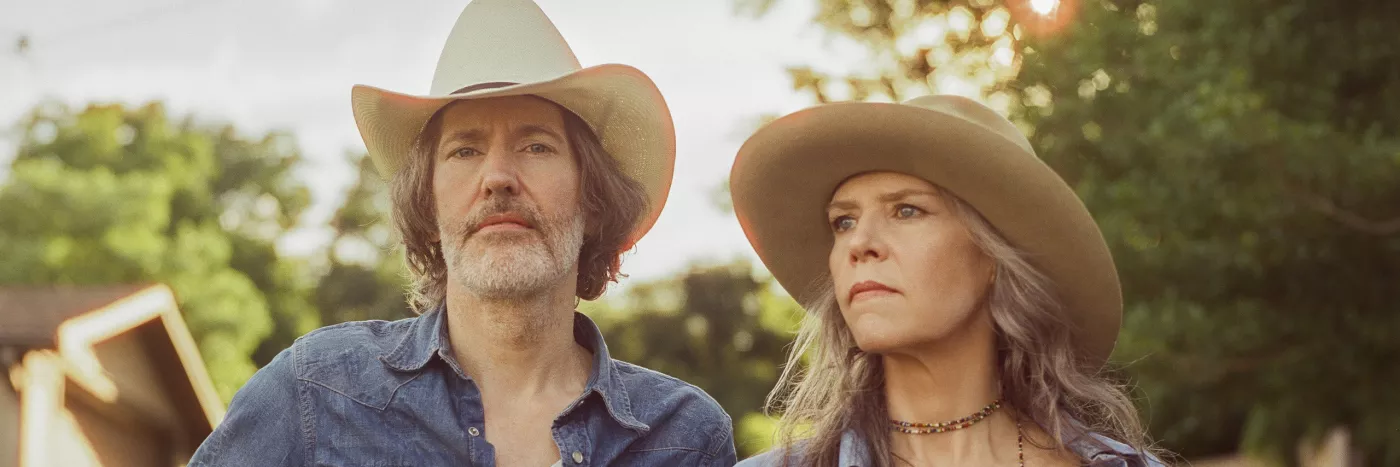 A man holding a guitar and woman both in cowboy hats look pensive away from the camera exuding a country music vibe.