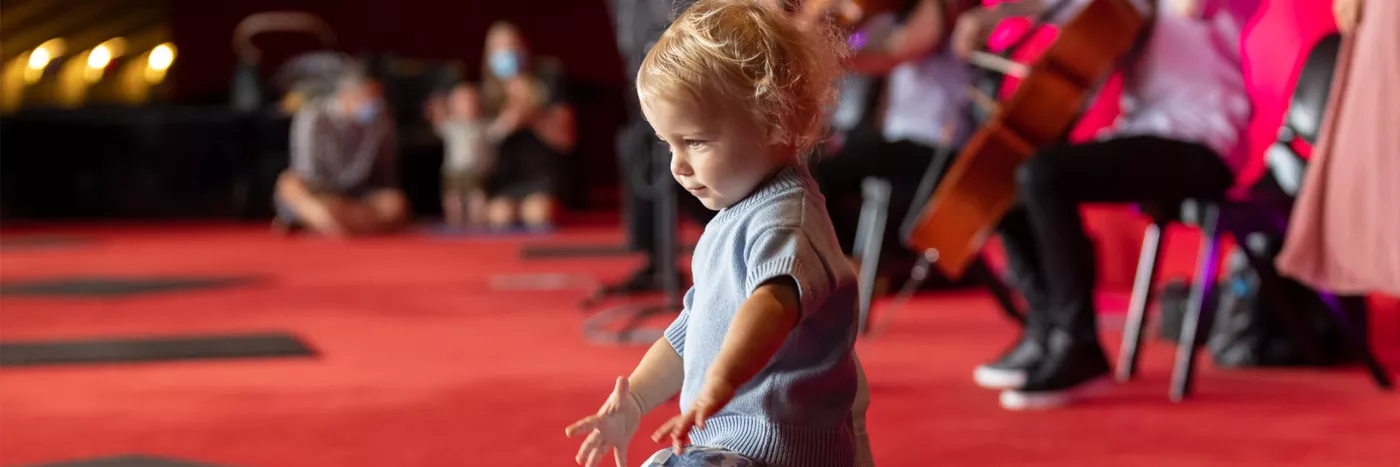 A toddler in a blue sweater and floral skirt dances on a red carpeted floor with people and musicians playing string instruments in the background.