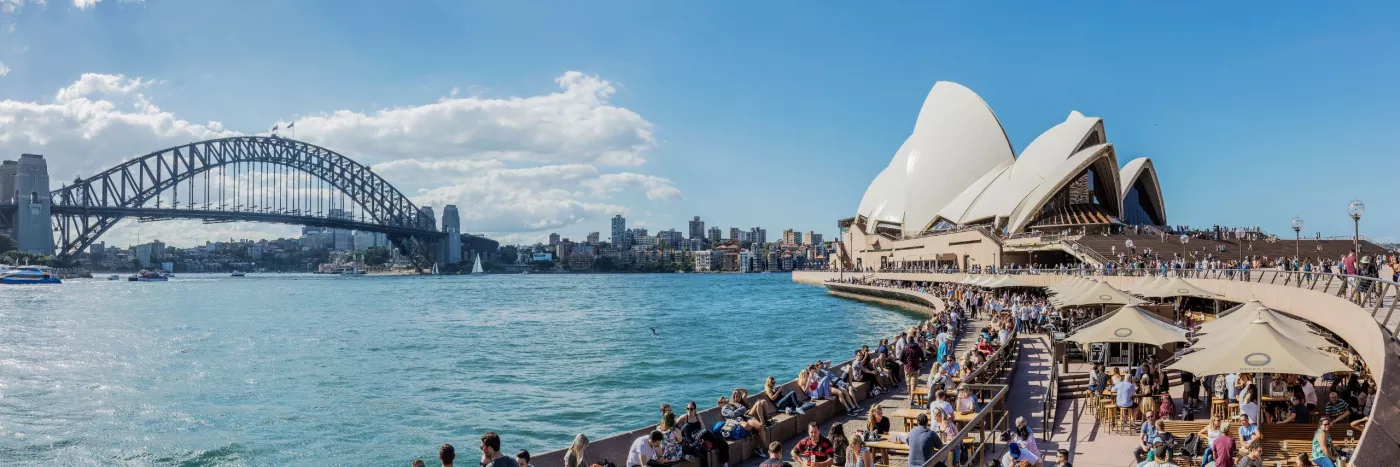 Panoramic view of Sydney Harbour with the Sydney Opera House on the right and Sydney Harbour Bridge on the left, with numerous people and waterfront restaurants in the foreground.