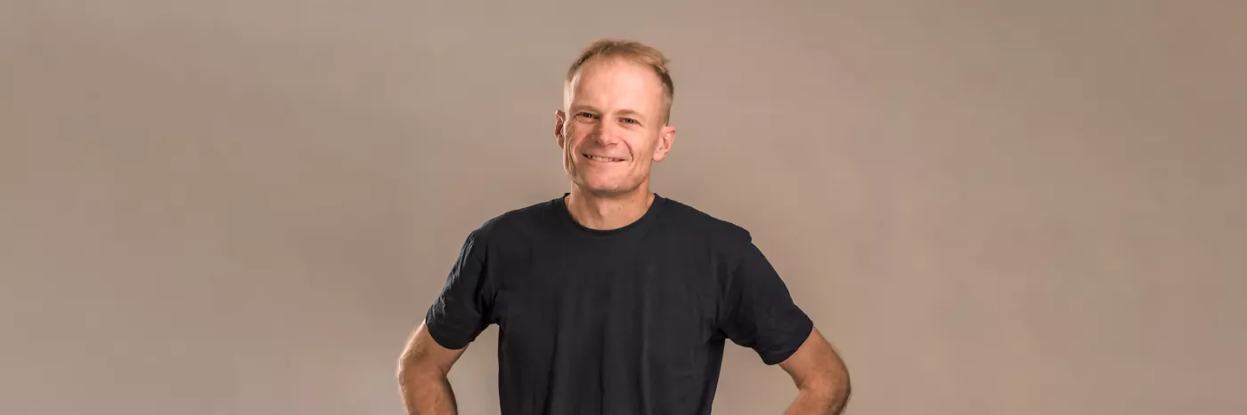 A man with short light brown hair smiles with his hands on his hips. He wears a black t-shirt.