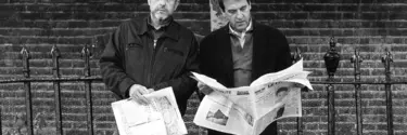 Two men with newspapers, standing in front of a brick wall and iron gates.