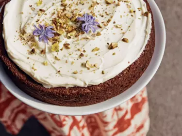 A carrot cake with icing and three purple flowers on it on a white plate being held by a person who is out of frame.